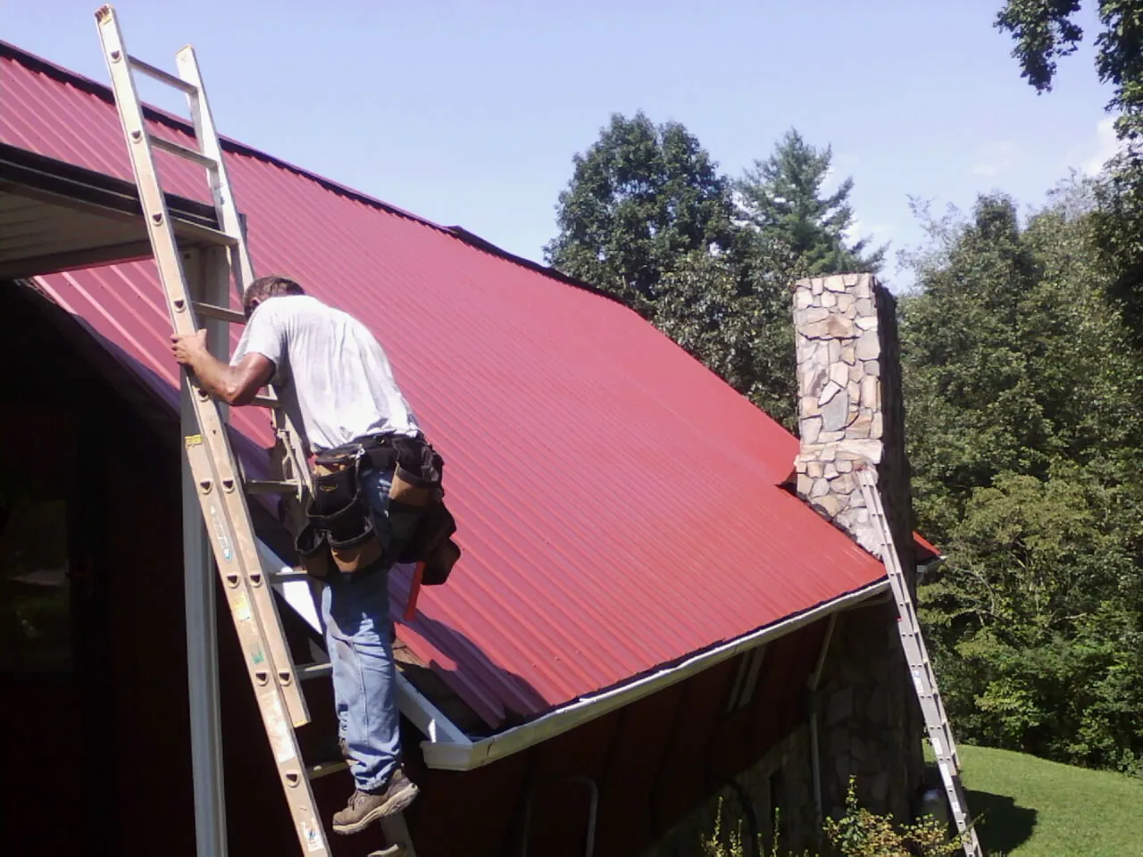 Roofer on a ladder beside a red metal roof and stone chimney — Soffit & Fascia Repair by Roofing Brothers in Powells Point